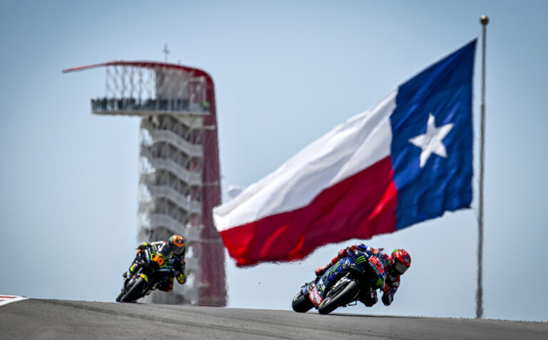 Two COTA MotoGP Racers on Track with Flag in Background