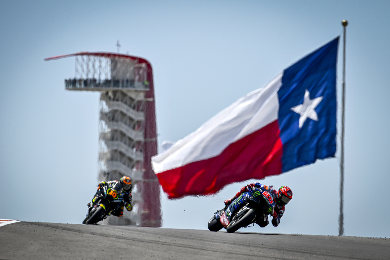 Two COTA MotoGP Racers on Track with Flag in Background